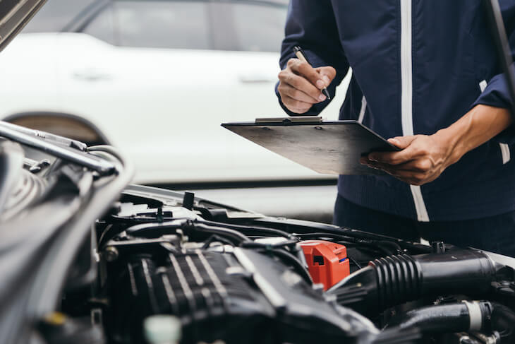 mechanic performs car inspection looking at the engine of a car with clip board in hand